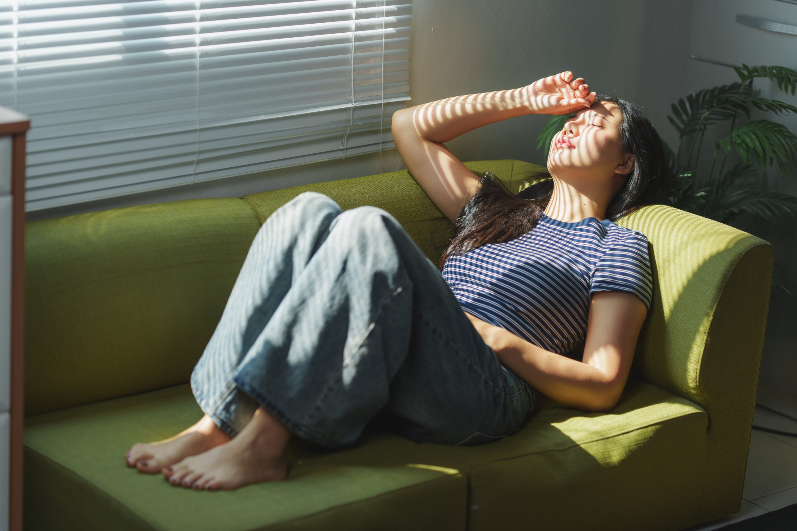 Young woman is lying on a green sofa with her hand on her forehead, enjoying the sunlight filtering through the blinds, creating a peaceful and relaxing atmosphere